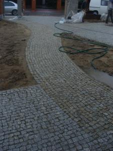 Ułożona ścieżka z kostki granitowej o nieregularnym kształcie, z widocznym fragmentem obszaru przygotowanego pod dalsze układanie, wężykiem i budynkiem w tle.